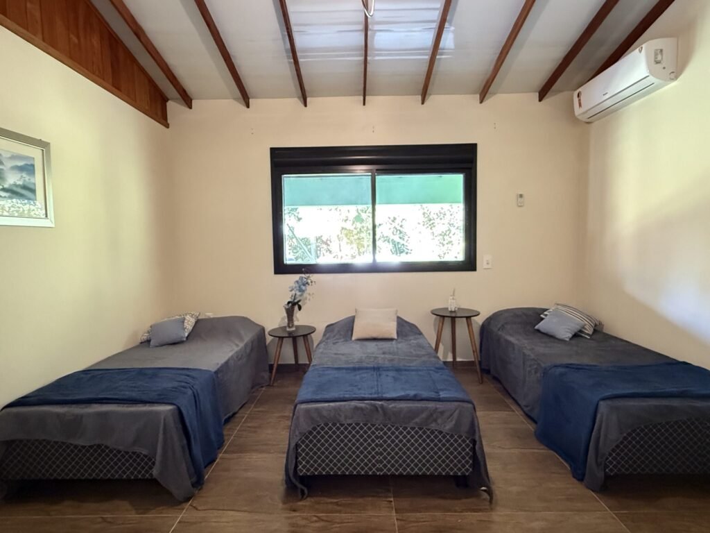 Quarto com três camas, duas mesas de cabeceira, e janela ensolarada