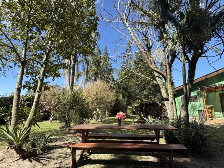 Banco com flores em área verde arborizada, com casa ao fundo, em dia ensolarado